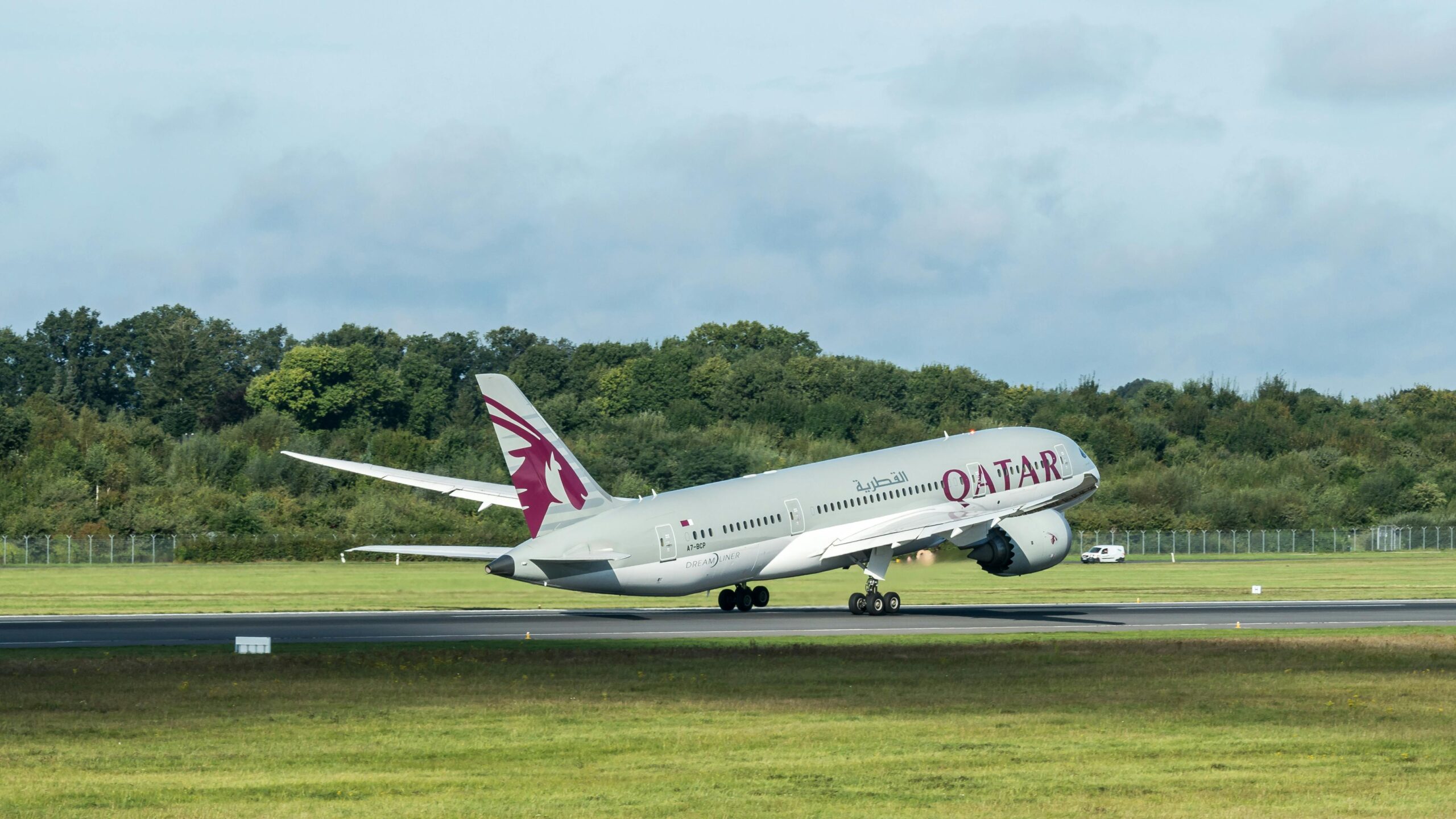 Qatar Airways aircraft takes off from a grassy runway surrounded by trees on a clear day.